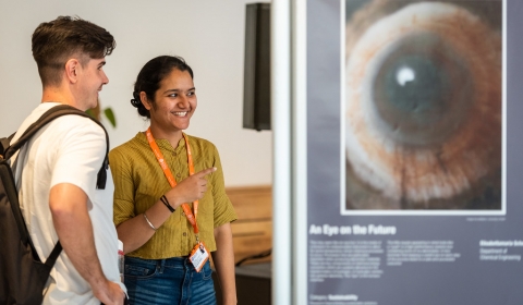 A caucasian male and South Asian female stand next to a exhibition panel in discussion