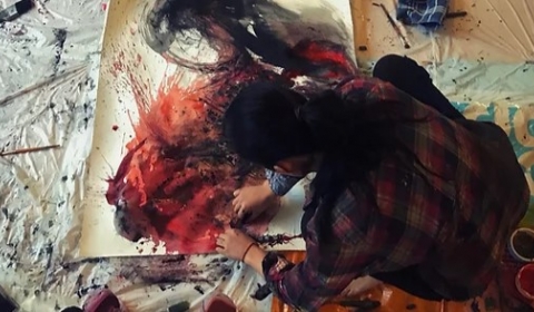An female Latin artist seated on the floor painting on a canvas