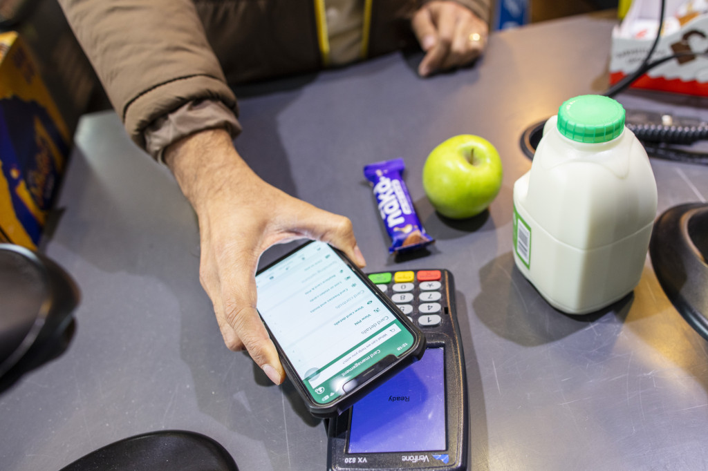 Photograph of grocery items at a supermarket checkout, with a customer paying using a smartphone. The image highlights food costs, household budgeting, and affordable access to food during the cost of living crisis.