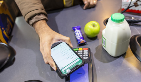 Photograph of grocery items at a supermarket checkout, with a customer paying using a smartphone. The image highlights food costs, household budgeting, and affordable access to food during the cost of living crisis.