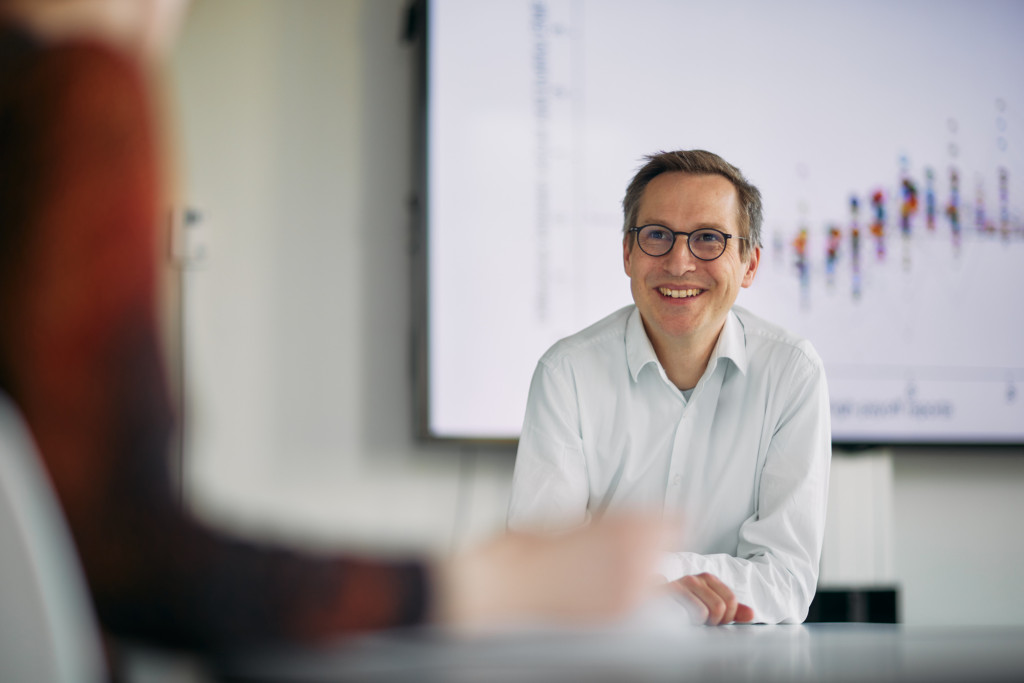 Photograph of Professor Johannes Abeler, a white male in a white shirt, sitting in front of a screen showing a blurred graph