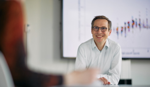 Photograph of Professor Johannes Abeler, a white male in a white shirt, sitting in front of a screen showing a blurred graph