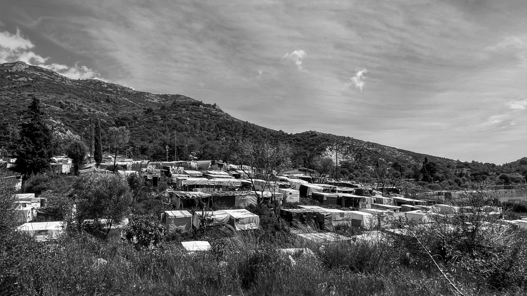 Black and  landscape image Samos. Grass, trees, a mountain and blue sky frame a large camp made up of huts.