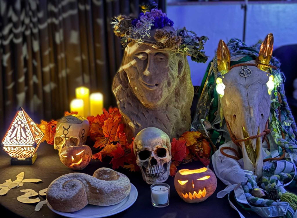 Photograph of an arrangement of item placed a surface in a dark room. Items include candles, carved pumpkin, offerings, skulls and sculpture of a face.