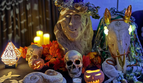 Photograph of an arrangement of item placed a surface in a dark room. Items include candles, carved pumpkin, offerings, skulls and sculpture of a face.
