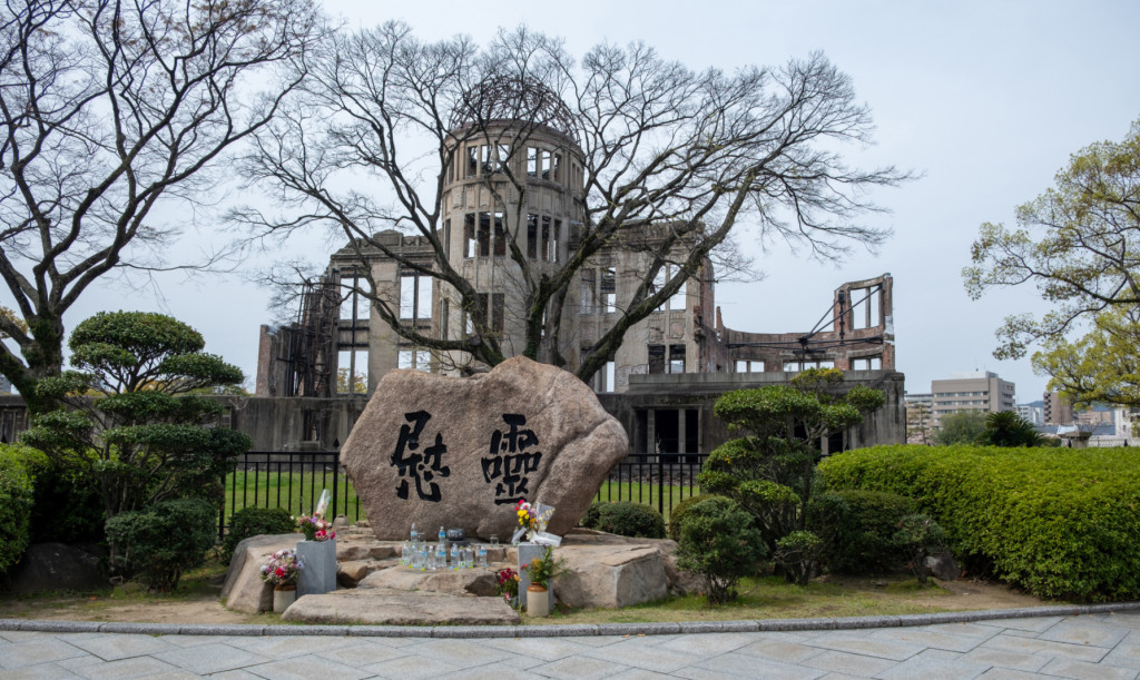 A memorial stone with Japanese characters sits in the foreground, with the skeletal remains of the Hiroshima Peace Memorial (Genbaku Dome) in the background. Bare trees and trimmed bushes surround the monument.