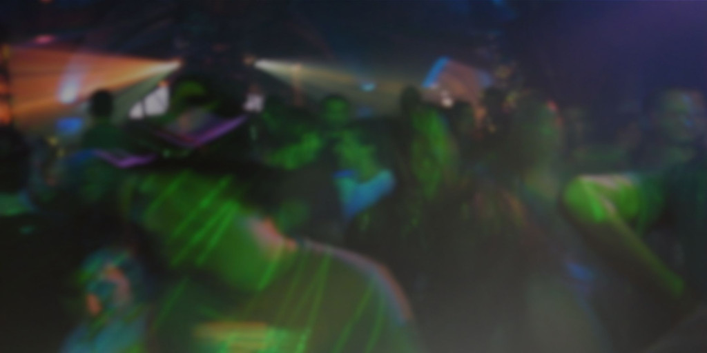 Photograph of a crowded nightclub scene with people dancing beneath intense coloured lighting. Green, blue, purple and orange beams create blurred motion.