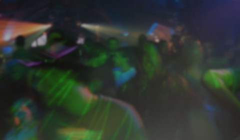 Photograph of a crowded nightclub scene with people dancing beneath intense coloured lighting. Green, blue, purple and orange beams create blurred motion.