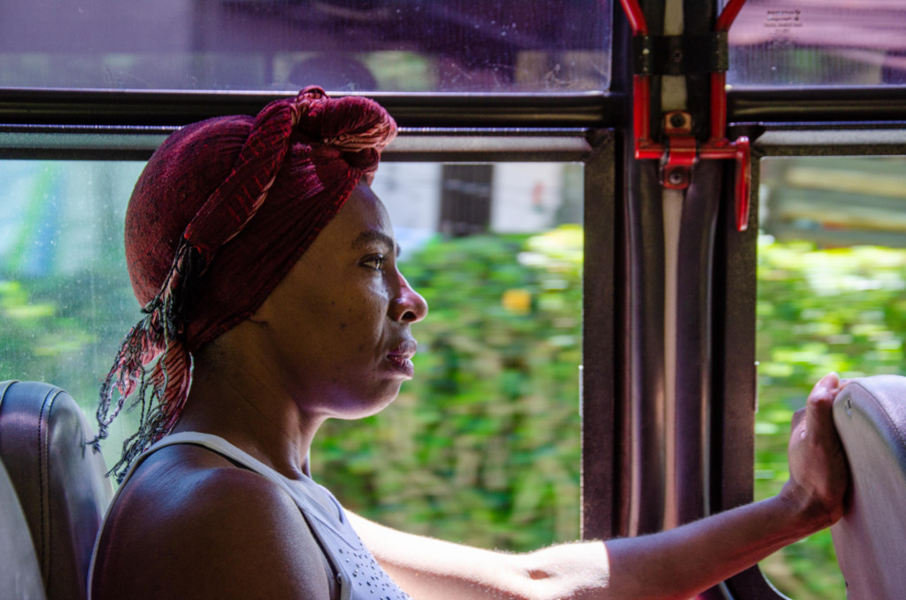 A photograph of a black woman with a red scarf tied over her head, sitting on a bus, looking forward with a serious expression.
