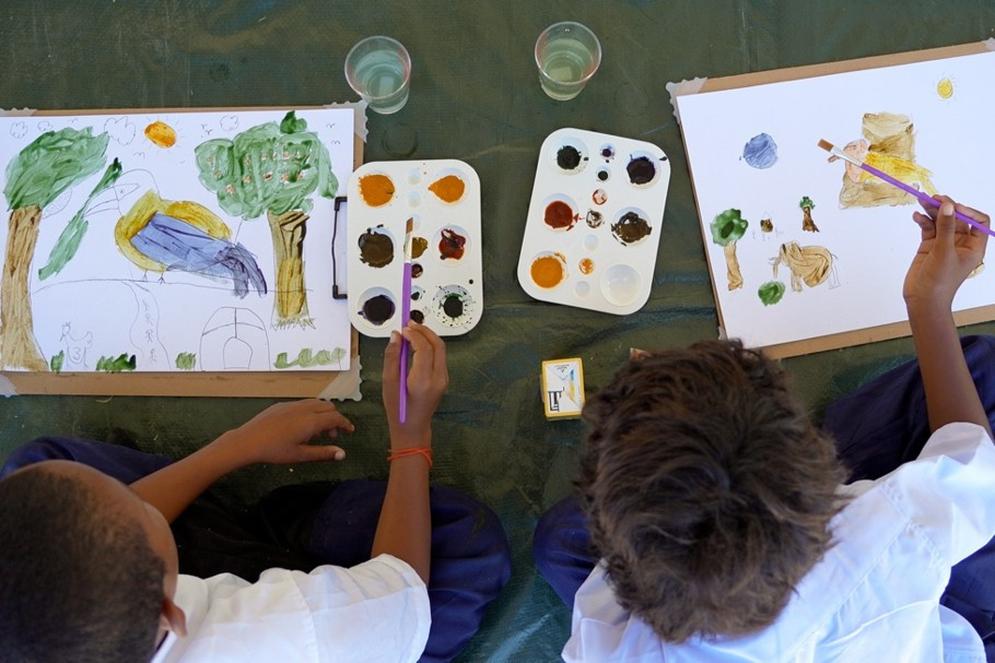 Two children viewed from above creating paintings of local plants and animals using natural watercolours, with paint palettes and brushes arranged between their drawings.