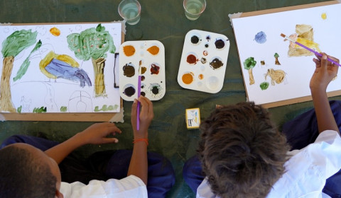 Two children viewed from above creating paintings of local plants and animals using natural watercolours, with paint palettes and brushes arranged between their drawings.