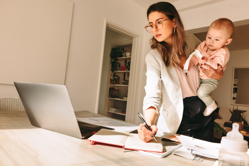 A mother multitasks, holding her child on one hip as she manages a computer and scattered workbooks.
