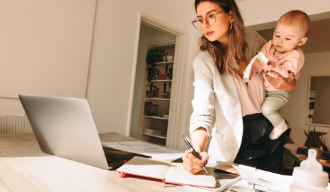 A mother multitasks, holding her child on one hip as she manages a computer and scattered workbooks.