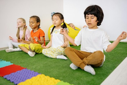 A diverse group of four children sit in a white room on artificial grass. Each of them sits with crossed legs and eyes closed in a lotus position.