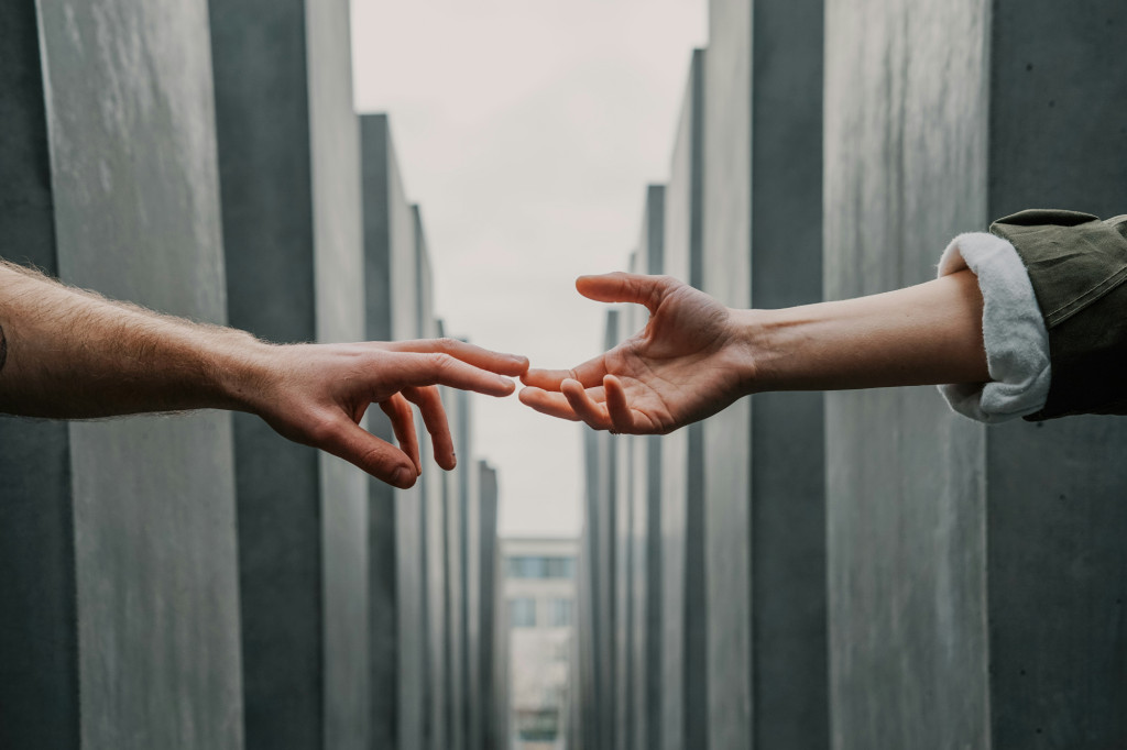 two hands reach out to each other, fingertips just making contact, reminiscent of 'The Creation of Adam' painting by Michelangelo, set against a stark background of concrete structures