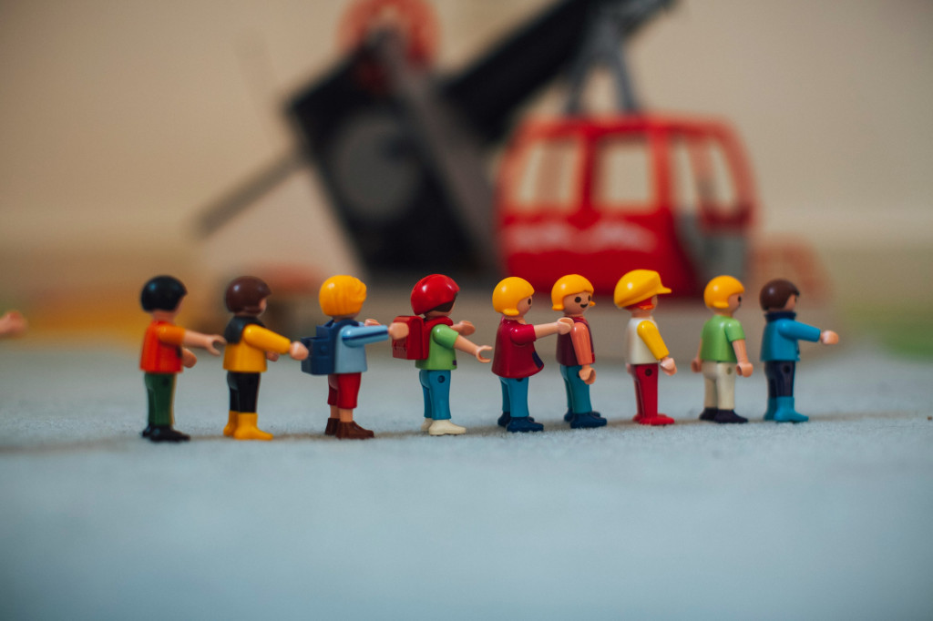 Photo of miniature toy lego-style figurines in the foreground, walking in a line. The background is out of focus and shows of a piece of machinery. Some of the figures wear hard hats and carry backpacks, one's head is turned smiling at us.