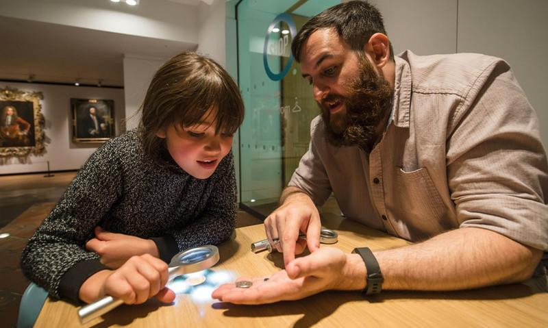 A child examines a coin with a magnifying glass while a bearded man holds another coin in his hand at a museum.