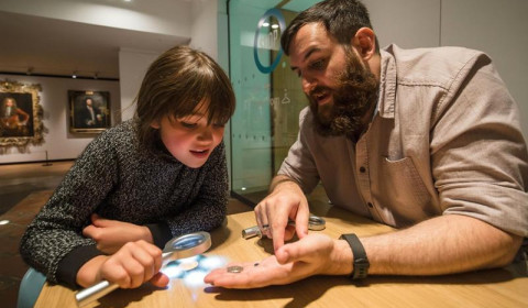 A child examines a coin with a magnifying glass while a bearded man holds another coin in his hand at a museum.