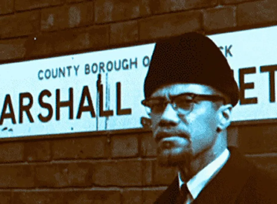 The image shows civil rights activist Malcolm X standing in front of a street sign for Marshall Street during his visit to Smethwick, UK, in February 1965.
