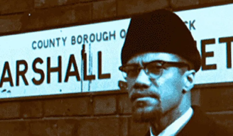 The image shows civil rights activist Malcolm X standing in front of a street sign for Marshall Street during his visit to Smethwick, UK, in February 1965.