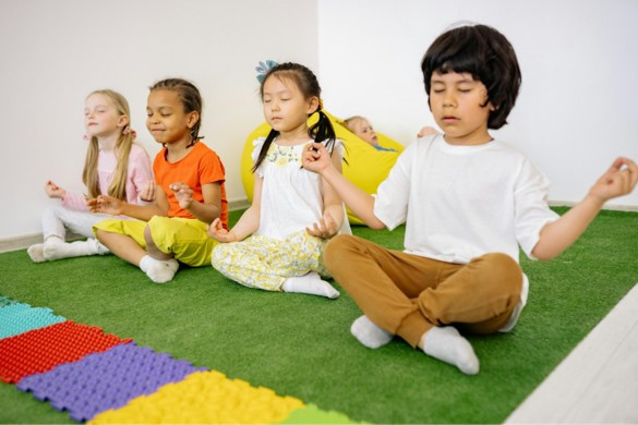 Four children sit in a row on a grass-like mat. They sit with their legs cross, eyes closed, and fingers pressed to thumbs.