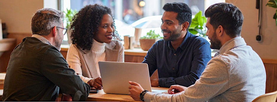 A photo of four people sitting round a table smiling and conversing, with a laptop open in front of them.