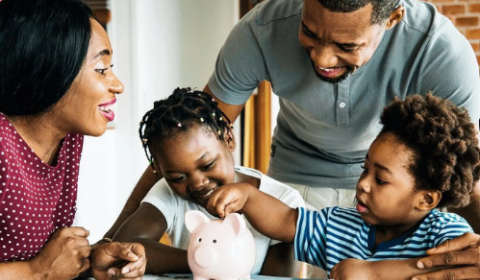 Family gathered around a table as two children put coins into a pink piggy bank.