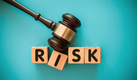 The word RISK, spelt out from left to right in wooden building blocks with one letter on each building block. The building blocks are being hit with a legal hammer, placed in front of a plain blue background