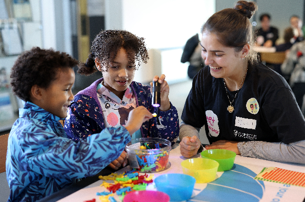 Two children and a woman at a table smiling, while the children interact with colourful toy figures, a torch, and brightly coloured plastic bowls.