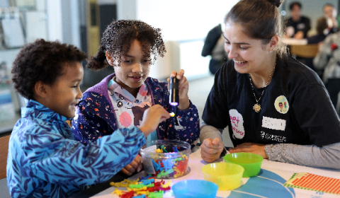 Two children and a woman at a table smiling, while the children interact with colourful toy figures, a torch, and brightly coloured plastic bowls.