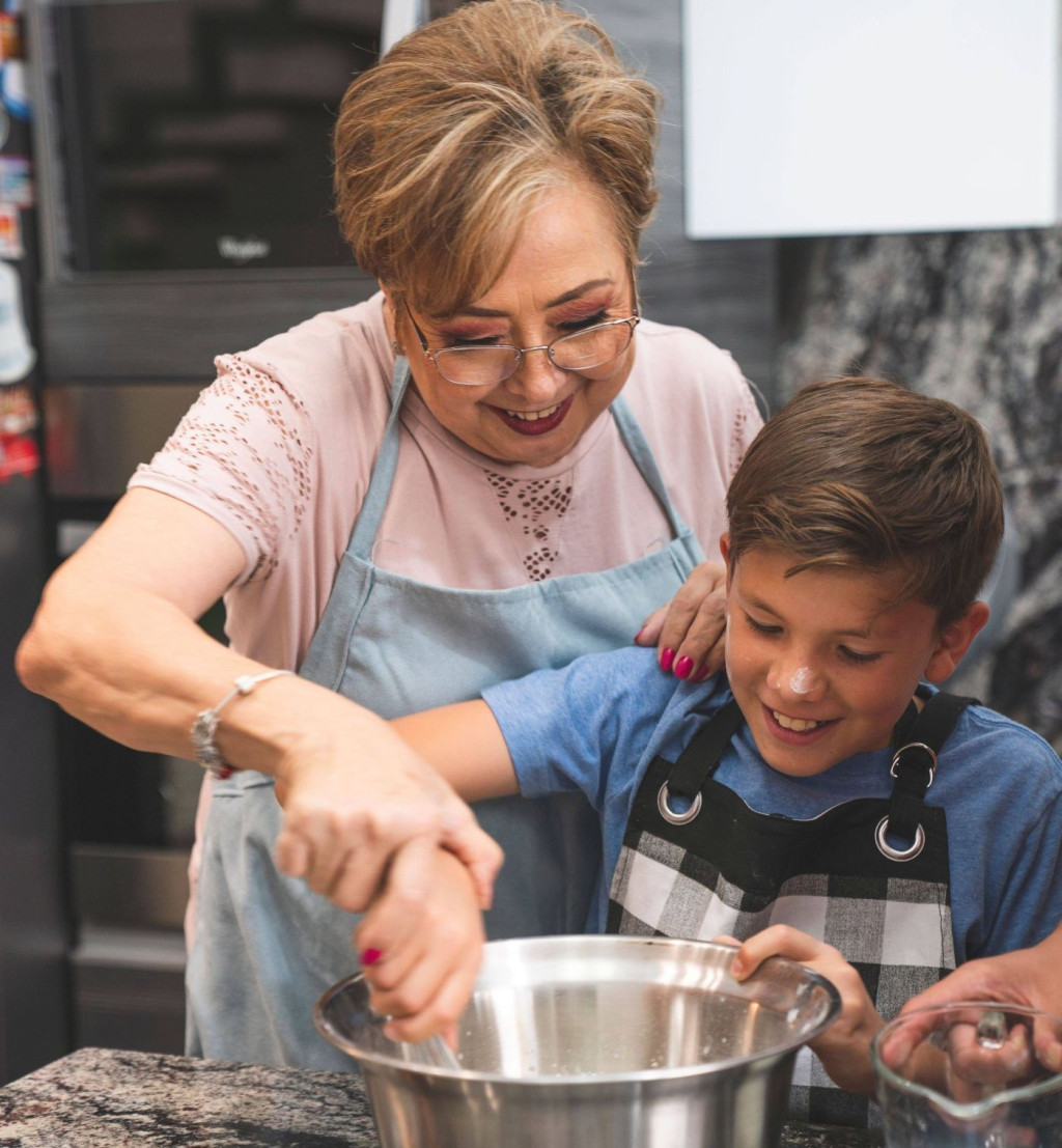 Image of a grandmother cooking with grandson, smiling as she is helping grandson to stir the pot.