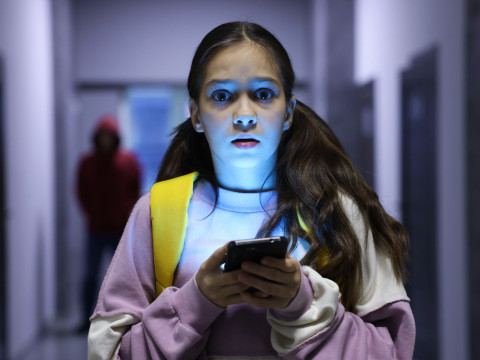 The picture shows a young girl standing in a dimly lit hallway while looking at her smartphone and a hooded preditor is in the background.