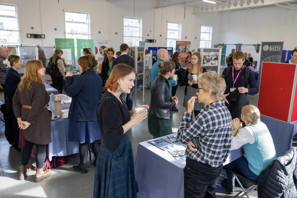 A busy room where multiple groups of people talk around tables and exhibition stands