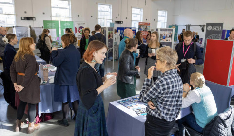 A busy room where multiple groups of people talk around tables and exhibition stands