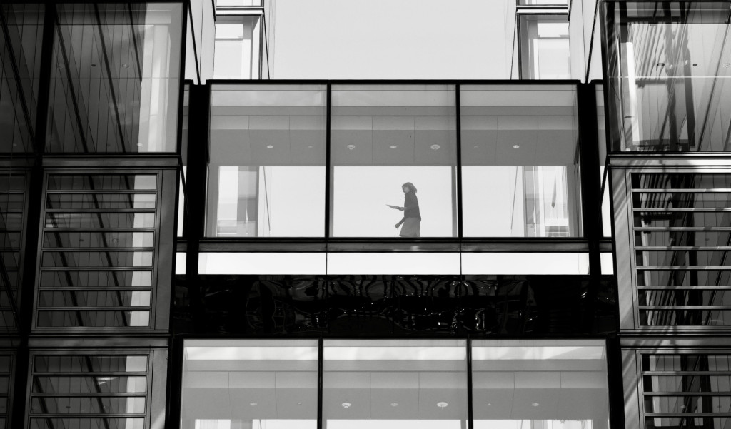 Black and white photo of a single woman walking across an enclosed platform between two building blocks
