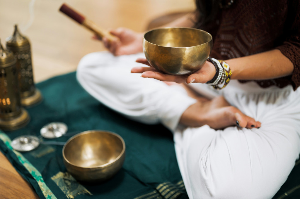 Woman wearing white trousers and a black long sleeved top is sat cross legged on a green mat.  In she is holding a small sound bowl in her left hand.