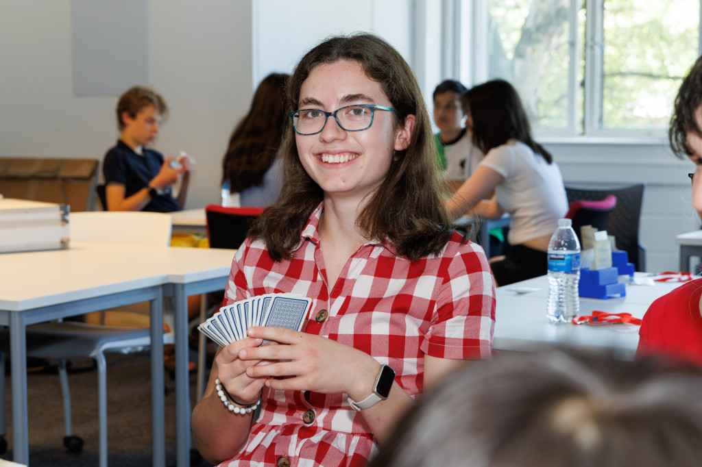 An image of a girl with shoulder length brown hair and blue glasses wearing a red checked shirt. She is smiling and holding cards in her hand.