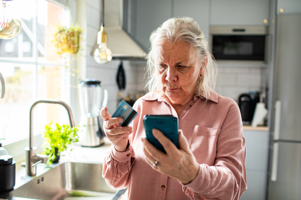 The picture shows an older woman standing in a bright, modern kitchen. She is holding a smartphone in one hand and a payment card in the other, looking closely at the phone with a serious, concentrated expression.