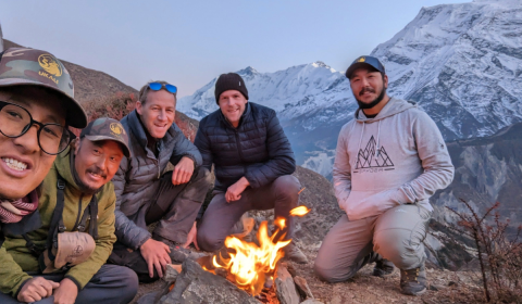 Photograph of a group of individuals around a camp fire in the mountains, with a snow covered mountain in the background