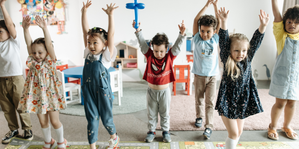Young children standing in a line in a room with toys in the background. The children have their arms up in the air.