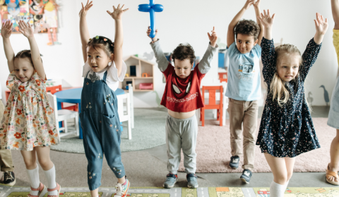 Young children standing in a line in a room with toys in the background. The children have their arms up in the air.