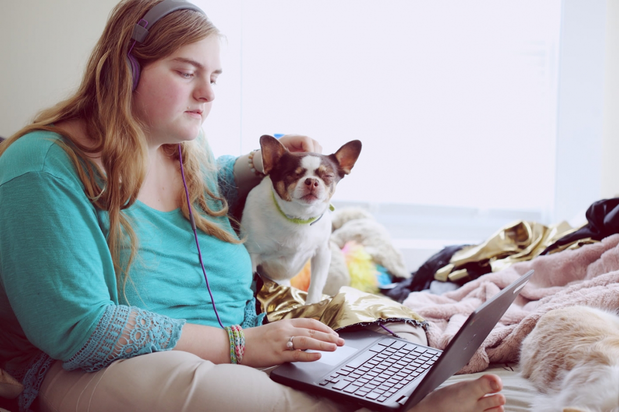 A photo of a young woman with blonde hair and a green top, sat with a small brown and white dog and surrounded by blankets. The woman is using her laptop and wearing headphones