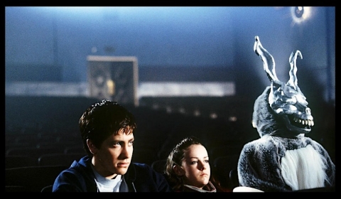 A frame from the film donnie darko with a man a women in a theatre with a creepy rabbit sitting beside them