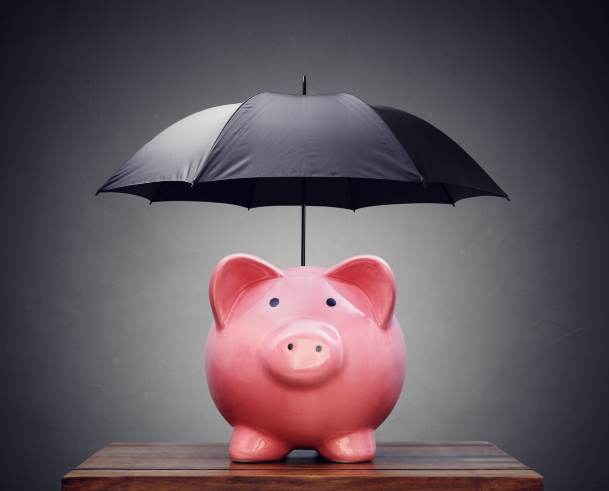 The picture shows a pink piggy bank sanding on a desk and covered by a black umbrella against a dark grey background.