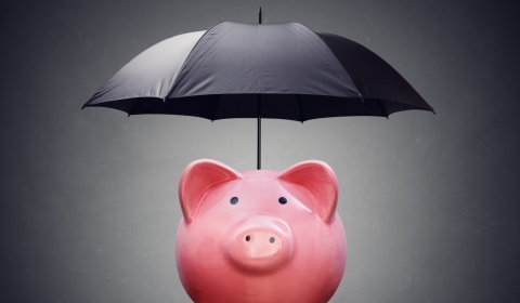 The picture shows a pink piggy bank sanding on a desk and covered by a black umbrella against a dark grey background.