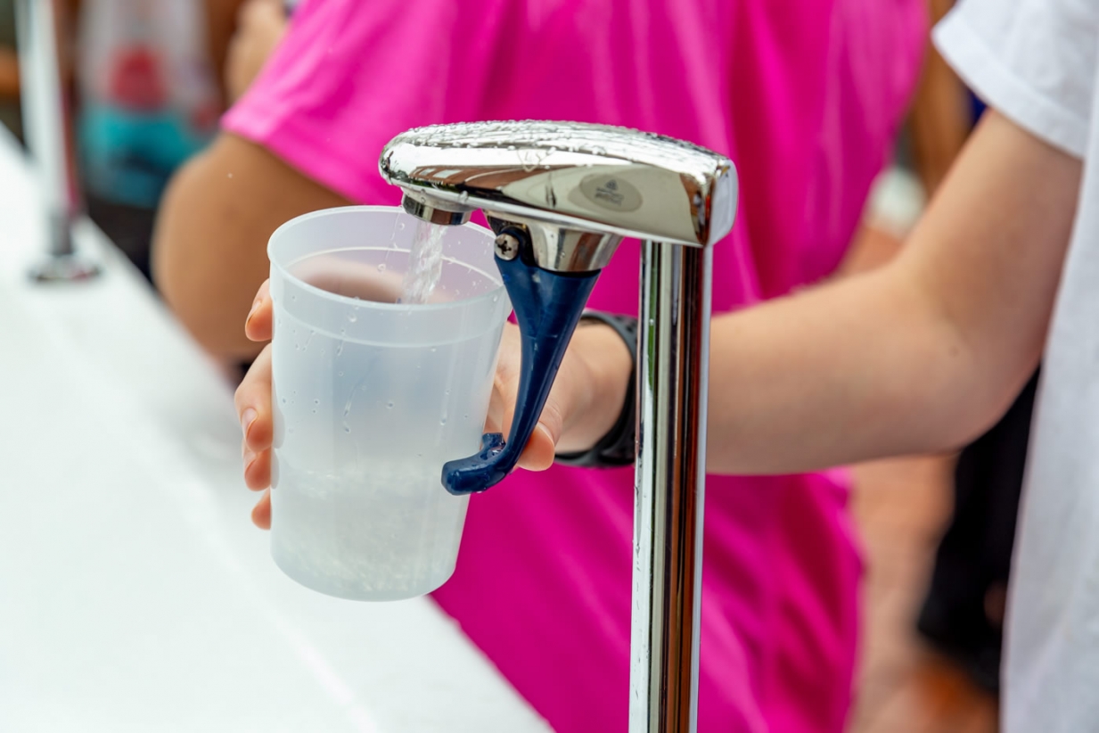 A person in pink top and a person holding a plastic cup underneath a water drinking fountain.
