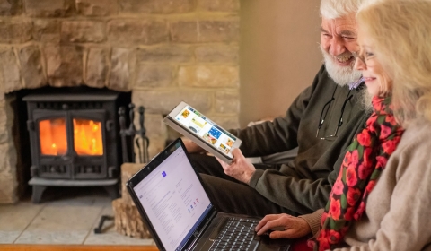 An older male and female sit on a sofa, holding a laptop and tablet. There is a cosy fireplace in the background.