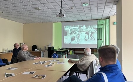 Photograph of men seated around a table watching footage of football on a large screen.