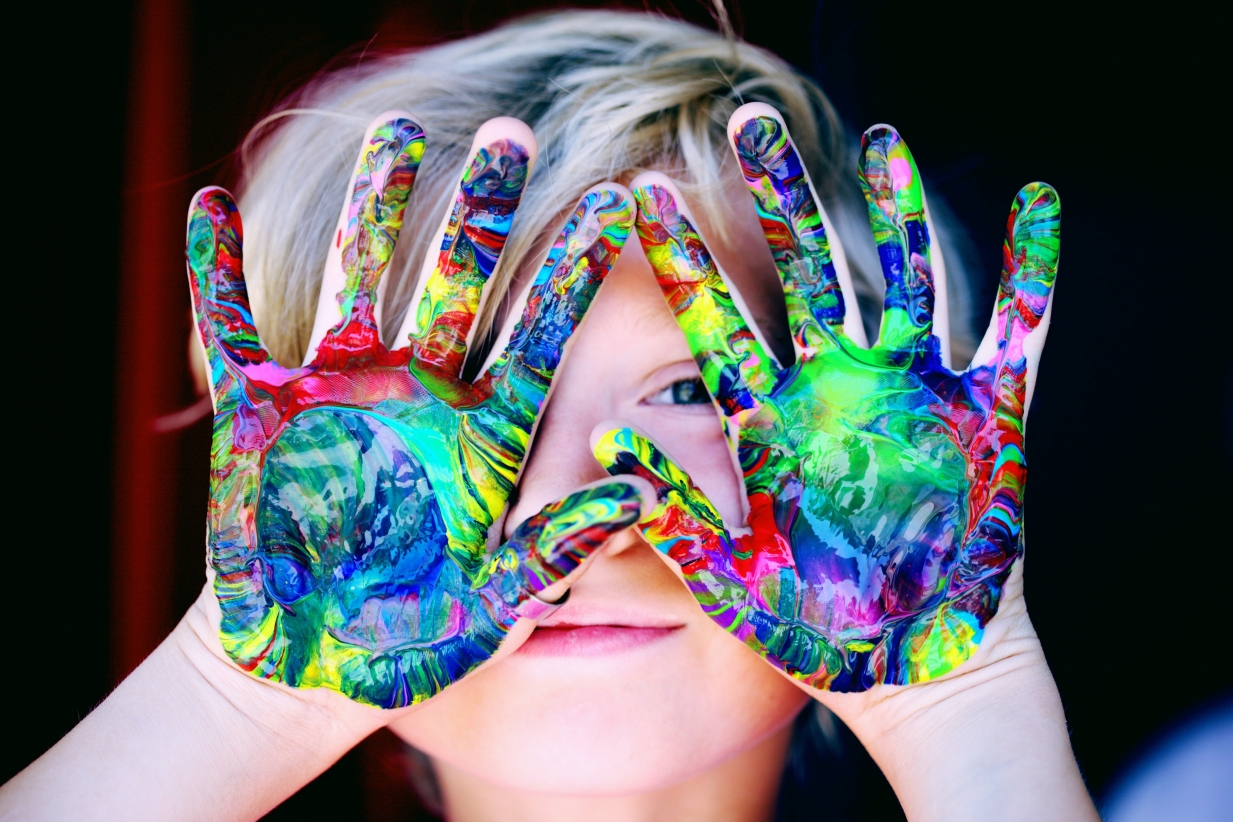 photo of a young smiling child holding their hands up palm outwards covering their face, their hands are covered in beautiful paint splashes