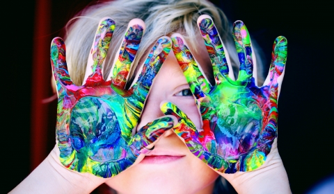 photo of a young smiling child holding their hands up palm outwards covering their face, their hands are covered in beautiful paint splashes